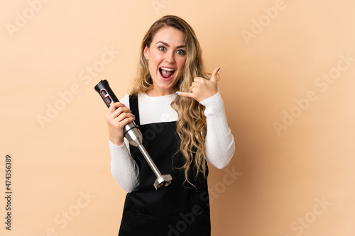 Young brazilian woman using hand blender isolated on beige background making phone gesture. Call me back sign