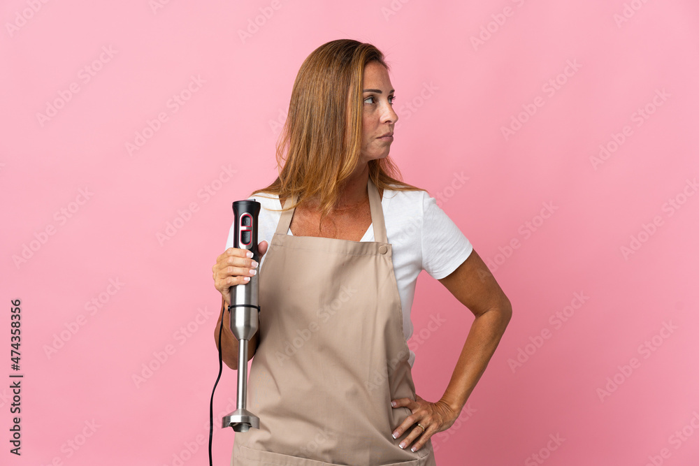 Middle age woman using hand blender isolated on pink background looking to the side Middle age woman using hand blender isolated on pink background looking to the side