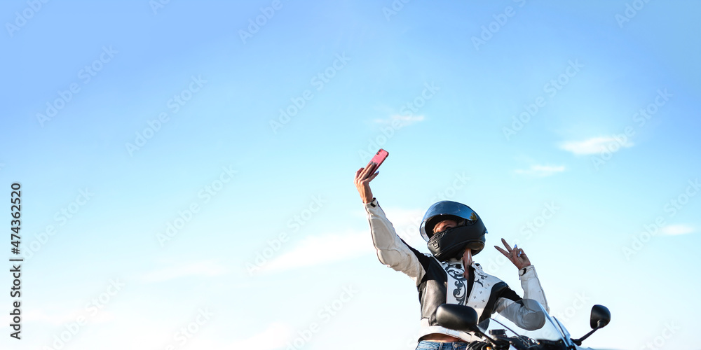 young woman biker with safety helmet taking a selfie with her smart phone making the gesture of the victory with her hand