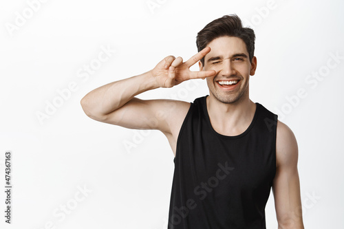 Happy healthy man in sport uniform showing peace sign and smiling. Sportsman express joy and enthusiasm after gym, white background © Cookie Studio