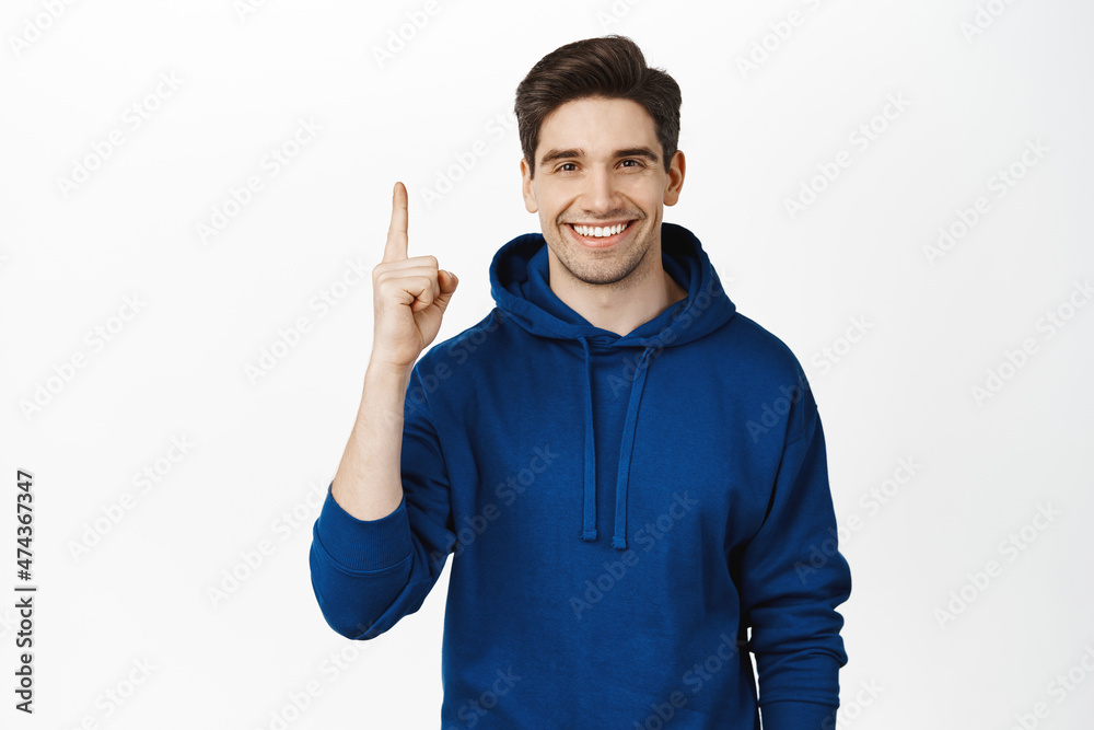 Fototapeta premium Smiling young man in hoodie, pointing finger up, showing advertisement, sale on top, standing over white background