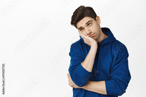 Bored and unamused brunette guy leaning on palm hand, looking away uninterested, stare at smth boring, standing over white background photo