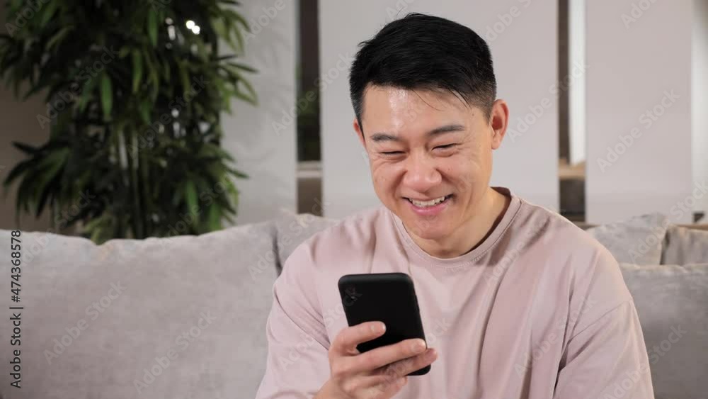 Smiling Asian Man Sitting on the Couch in Home Office Studio Using Smartphone, Man Reading Email Browsing Scrolling Tapping Through Internet, Social Online Networks Media. Smiling Asian Man Sitting on the Couch in Home Office Studio Using Smartphone, Man Reading Email Browsing Scrolling Tapping Through Internet, Social Online Networks Media.