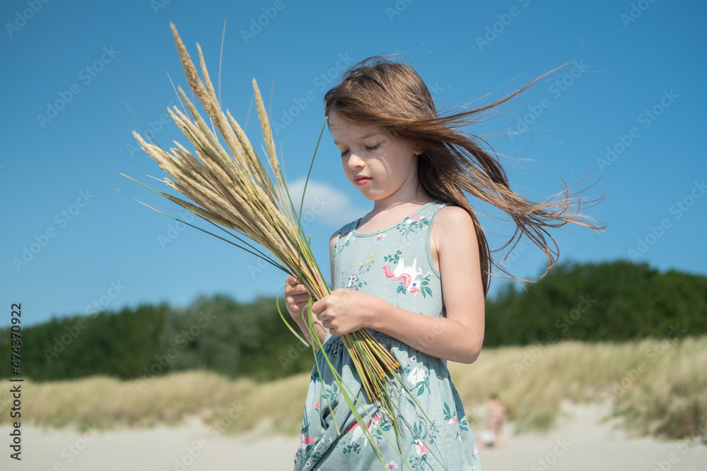 portrait of a small sad pensive girl in a dress, with long hair on the beach, with a bouquet of spikelets in her hand, summer time