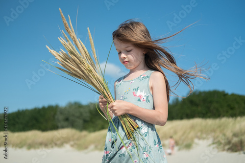 Obraz na płótnie portrait of a small sad pensive girl in a dress, with long hair on the beach, with a bouquet of spikelets in her hand, summer time w salonie