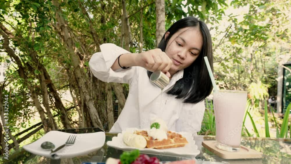 4k, A young Asian woman wearing a white shirt is pouring sweet syrup over a banana bread waffle on the table. ready to eat in the garden 4k, A young Asian woman wearing a white shirt is pouring sweet syrup over a banana bread waffle on the table. ready to eat in the garden