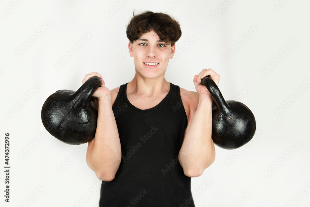 A young man, 16-18 years old, still a teenager with two heavy kettlebells on a light background, the guy is regularly engaged in weightlifting and looks strong and healthy