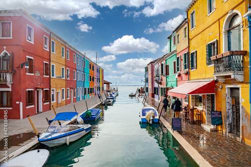 Burano Island, Venice, Italy. View of a canal in the town of Burano with the brightly colored houses famous all over the world. There are colored boats in the canal. Sunny blue sky with clouds.