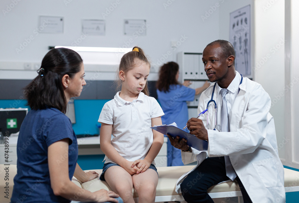 Obraz premium African american pediatrician doctor writing disease expertise of child on clipboard discussing medical treatment during clinical appointment in hospital office. Therapist man analyzing ill symptoms