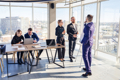 Confident businessman makes a presentation of a new project in the boardroom at a company meeting. Beautiful auditors talk with different partners about the business using a whiteboard and graphs.