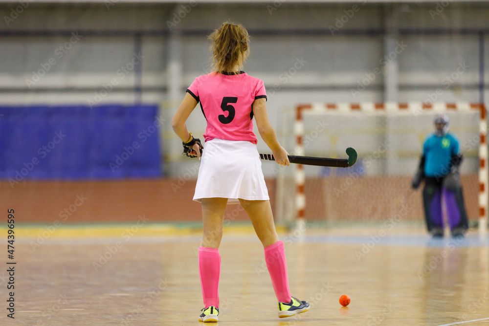 Young female indoor hockey player standing with the ball across goalkeeper.
