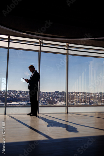 Rear view of a businessman looking out of a large window overlooking the city. He has a phone in his hands. Horizontal view. Selective focus