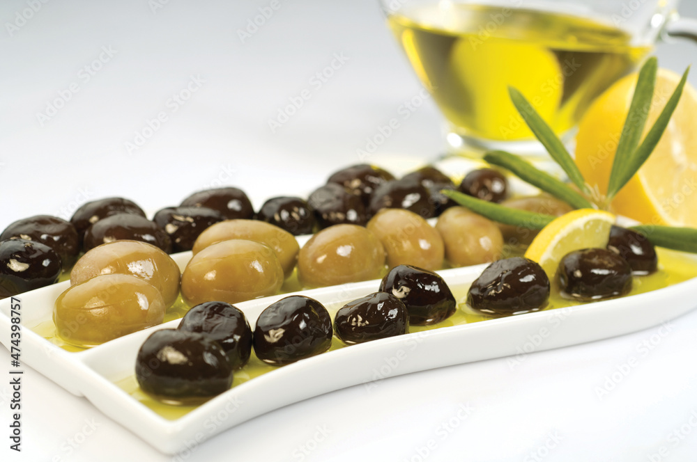 Glass bottle of homemade olive oil and olive tree branch, raw turkish green and black olive seeds and leaves on white table. Glass bottle of homemade olive oil and olive tree branch, raw turkish green and black olive seeds and leaves on white table.