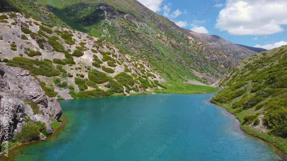 A drone flies over a turquoise mountain lake, two travelers next to a pond. the lake is surrounded by mountains and hills of konyon with small and large stones and various green vegetation. A drone flies over a turquoise mountain lake, two travelers next to a pond. the lake is surrounded by mountains and hills of konyon with small and large stones and various green vegetation.