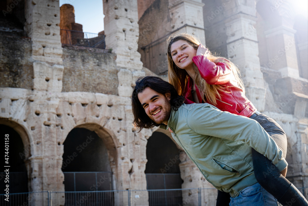 Young couple having fun traveling in Rome. Man carries his girlfriend on his back in front of the Colosseum.