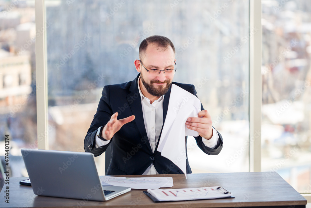 Fototapeta premium Adult male businessman, teacher, mentor is working on a new project in the office and is nervous. Sits at the table by the large window. He works on a laptop, business problems