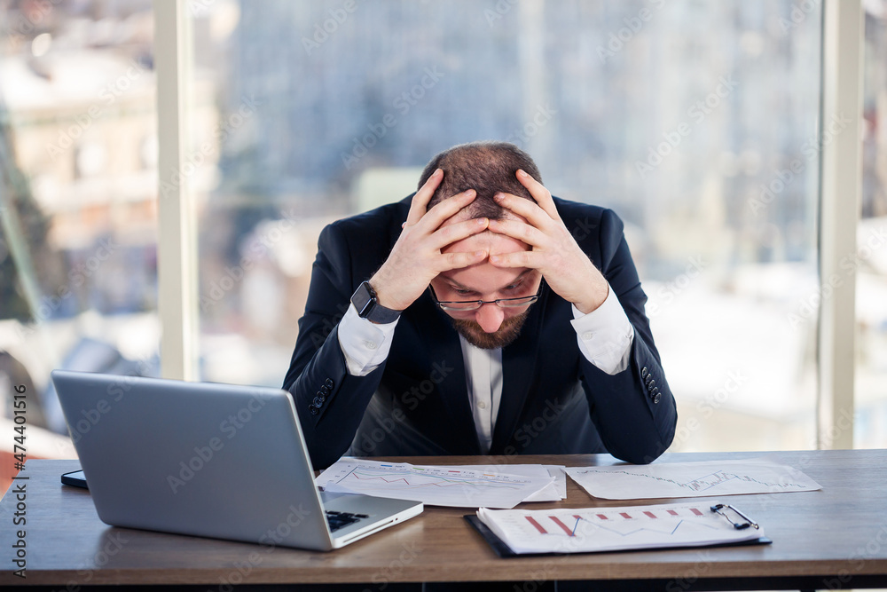 Adult male businessman, teacher, mentor is working on a new project in the office and is nervous. Sits at the table by the large window. He works on a laptop, business problems Adult male businessman, teacher, mentor is working on a new project in the office and is nervous. Sits at the table by the large window. He works on a laptop, business problems