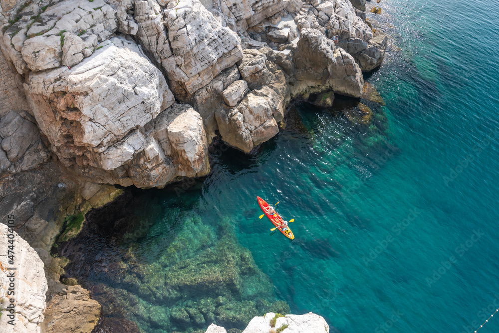 View from the rock cliffs of kayaker exploring the crystal clear Mediterranean waters of a cove off the coast of Dubrovnik, Croatia - obrazy, fototapety, plakaty 