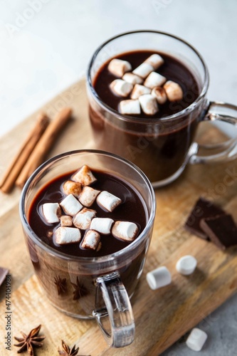 Two glass cups of hot chocolate with marshmallows and cinnamon sticks on wooden board and grey background. Winter food and drink concept. Selective focus.