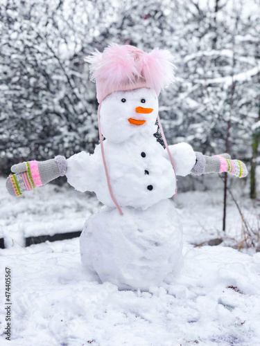 A snowman in a pink hat and gloves stands next to a snow-covered tree. Merry Christmas and Happy new year.
