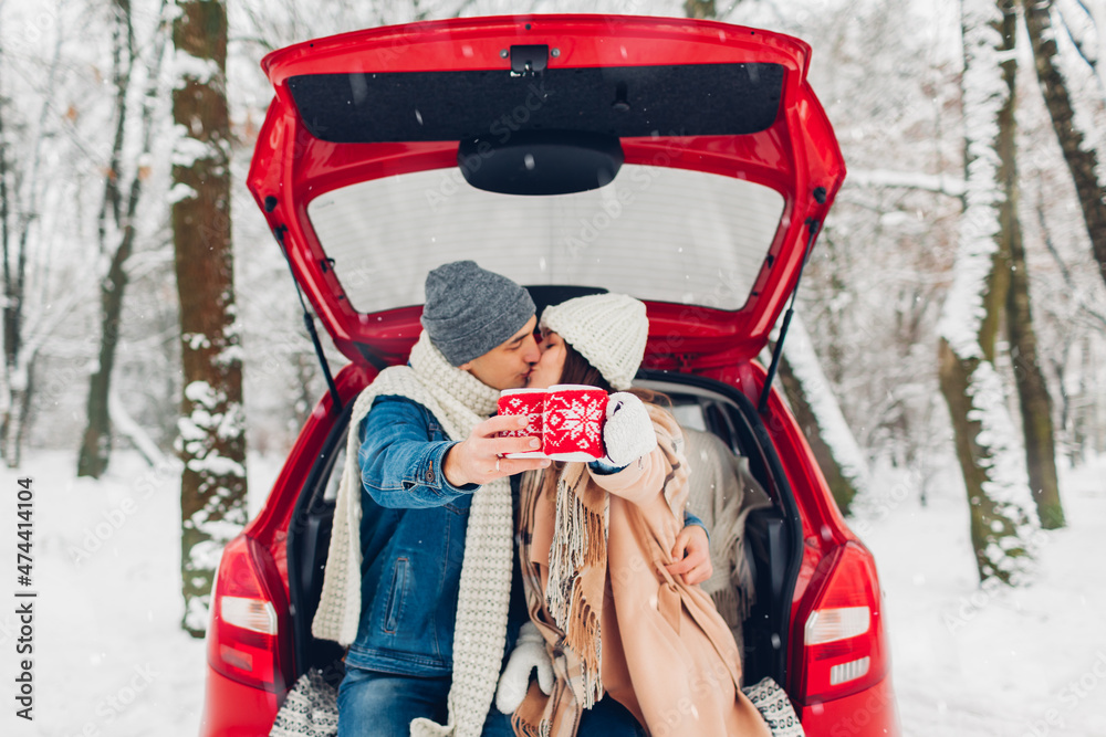 Kissing couple in love sitting in car trunk holding cups with tea in knitted Christmas cases in snowy winter forest.