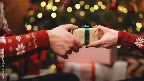 Close-up hands of man giving festive box with Christmas present to woman sitting at dinner feast table during holiday family party, decorating xmas tree and celebration lights, selective focus