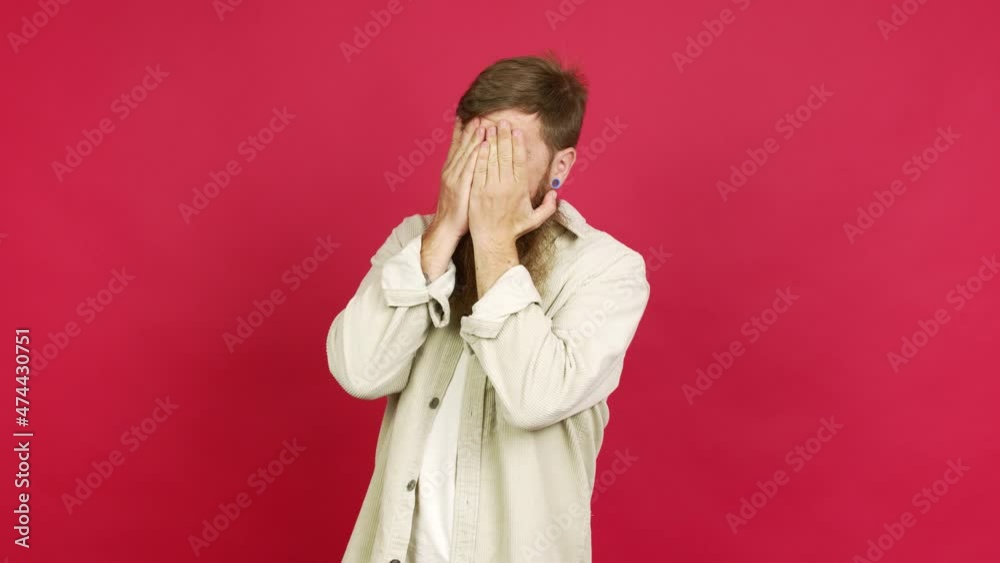 Redhead man with long beard covering eyes by hands and looking through the fingers over isolated background Redhead man with long beard covering eyes by hands and looking through the fingers over isolated background