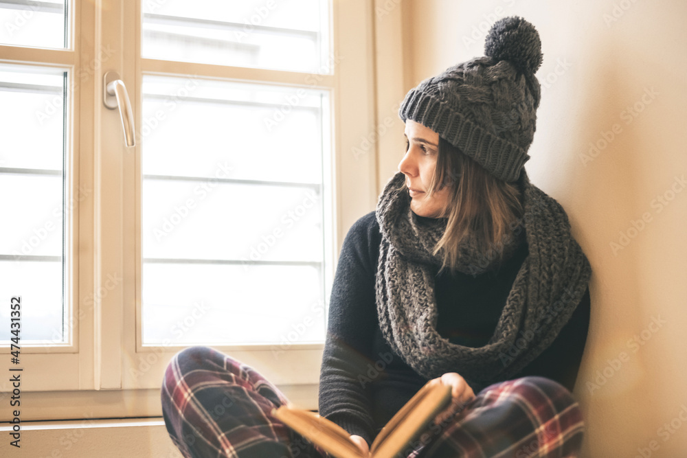 Young beautiful girl reading a book during cold winter with wool cap and scarf to keep warm. Woman sit read near the window. Female relaxing at home. Lifestyle lockdown carefree and education concept Young beautiful girl reading a book during cold winter with wool cap and scarf to keep warm. Woman sit read near the window. Female relaxing at home. Lifestyle lockdown carefree and education concept