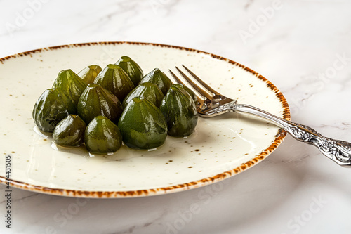 Close-up of conserved whole unripe figs and dessert fork on a white porcelain plate over marble surface. Traditional mediterranean jam confiture of green figs in syrup.