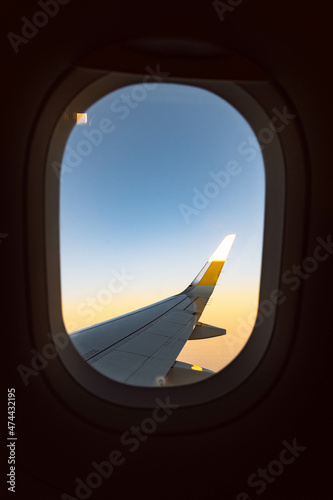 Fotografía en formato vertical de la ventanilla de un avión con el ala de fondo, con una franja amarilla al amanecer. photo
