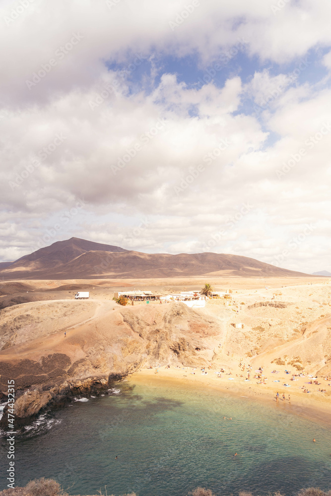 Fotografía vertical de un paisaje volcánico rocoso anaranjado con una playa y una montaña al fondo.