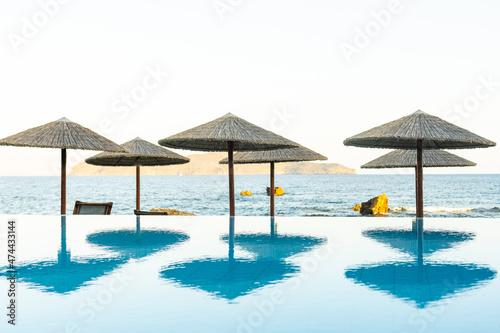 Deck chairs under sun umbrella between an infinity pool and the sea. Copy space provided on top.