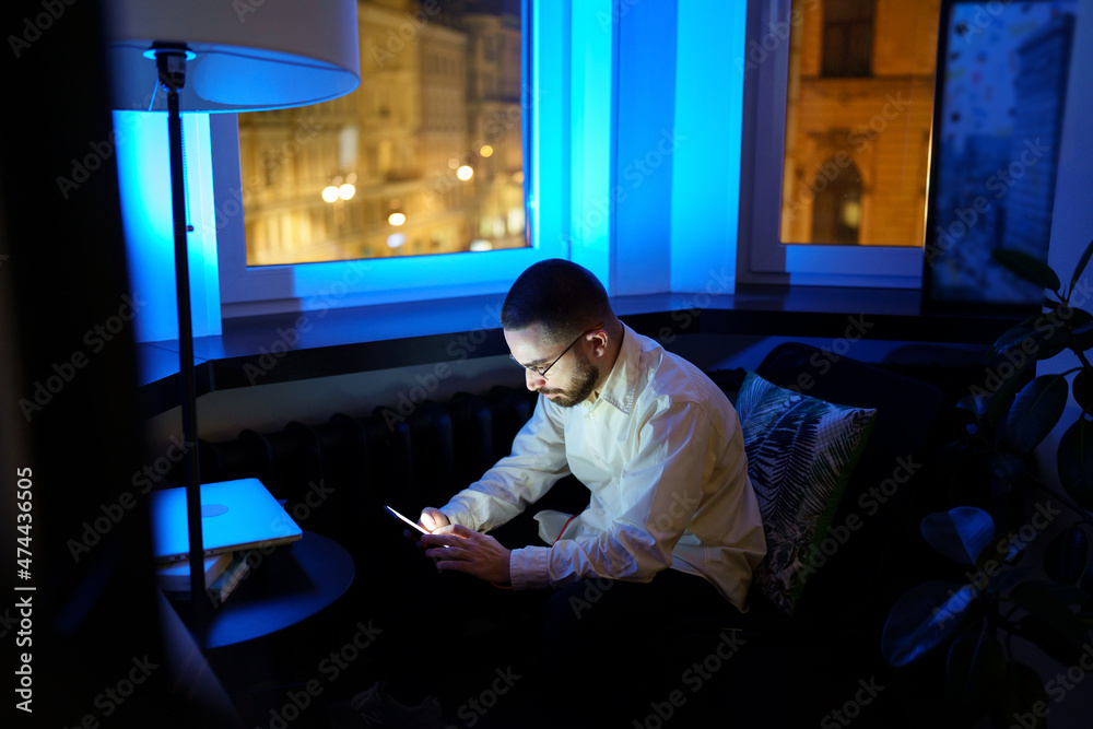 Tired young businessman texting home about staying at work overtime before deadline. Male programmer with smartphone in empty dark office. Stressed developer working at night. Overwork and tiredness