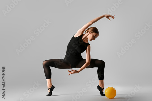 Beautiful young woman doing gymnastics with ball on grey background