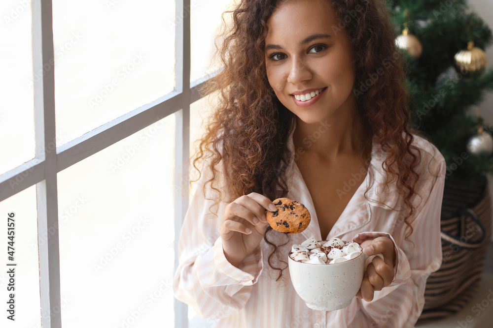 Young African-American woman with cookie and cup of hot cocoa near window on Christmas eve Young African-American woman with cookie and cup of hot cocoa near window on Christmas eve