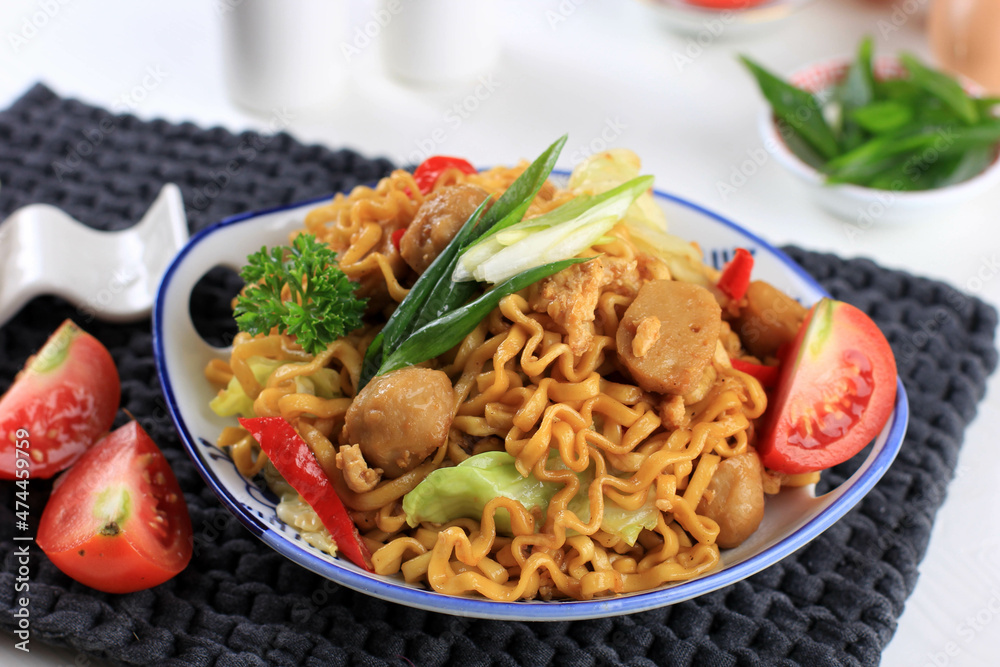 Mie goreng or Stir Fried Egg Noodles is Traditional Indonesian Food with Chicken, Egg, Tomato, Onion, Vegetable and Sweet Soy Sauce in White Plate Served on Wooden White Table. Top View. Mie goreng or Stir Fried Egg Noodles is Traditional Indonesian Food with Chicken, Egg, Tomato, Onion, Vegetable and Sweet Soy Sauce in White Plate Served on Wooden White Table. Top View.