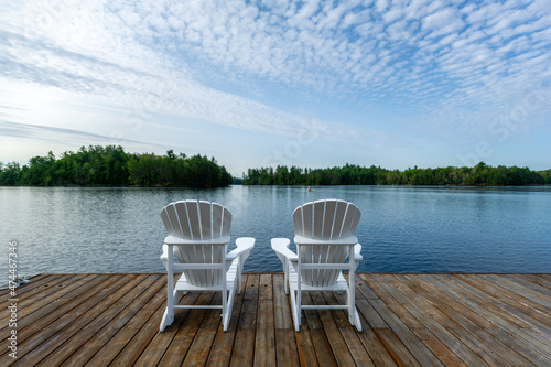 White Adirondack chair on a wooden dock facing the blue water of a lake in Ontario Canada. Cottages nestled among green trees are visible across the water. A single scull rower is passing.