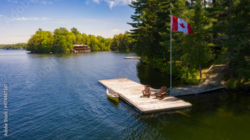 Two brown Adirondack chairs on a wooden pier with a yellow canoe. Across the calm water is a brown cottage nestled among green trees. Canada flag is waving on a pole. photo