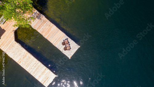 Aerial view of a cottage wooden pier on a lake in Muskoka, Ontario Canada. Two brown Adirondack chairs are visible on the dock facing the blue waters of the lake.