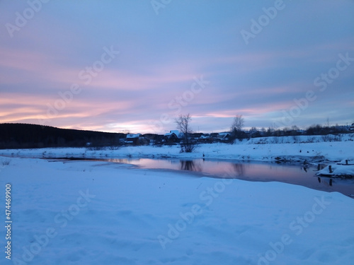 Winter snowy evening landscape in delicate pink and blue tones, a beautiful reflection of the sunset in the calm water of the river, a village and a forest in the distance