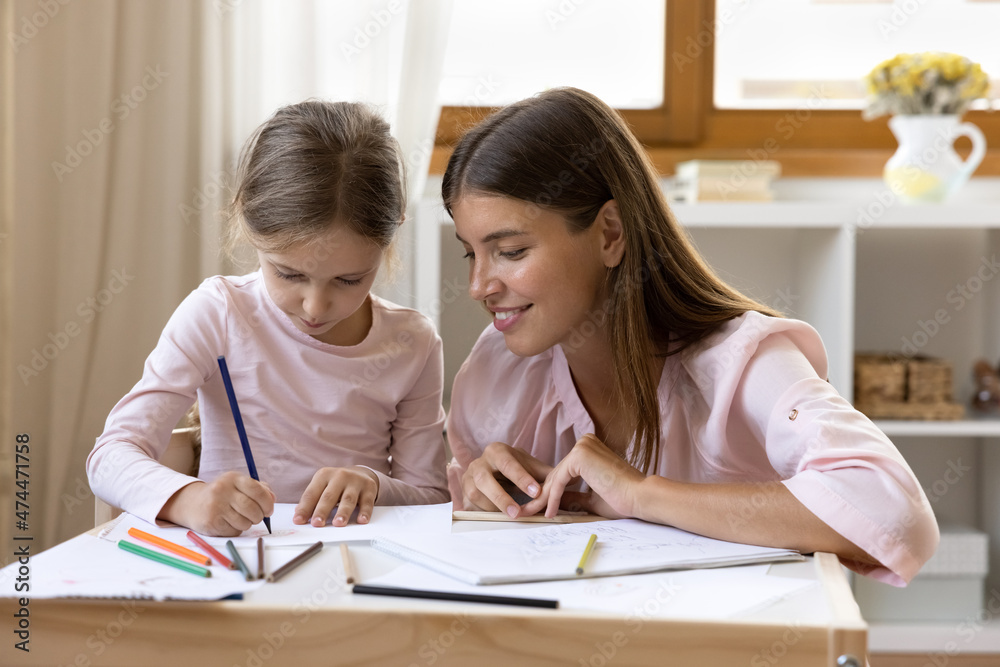 Happy preschool kids teacher watching focused little girl drawing in pencil in paper album. Mom helping cute daughter kid with school homework task. Childhood, child teaching concept Happy preschool kids teacher watching focused little girl drawing in pencil in paper album. Mom helping cute daughter kid with school homework task. Childhood, child teaching concept
