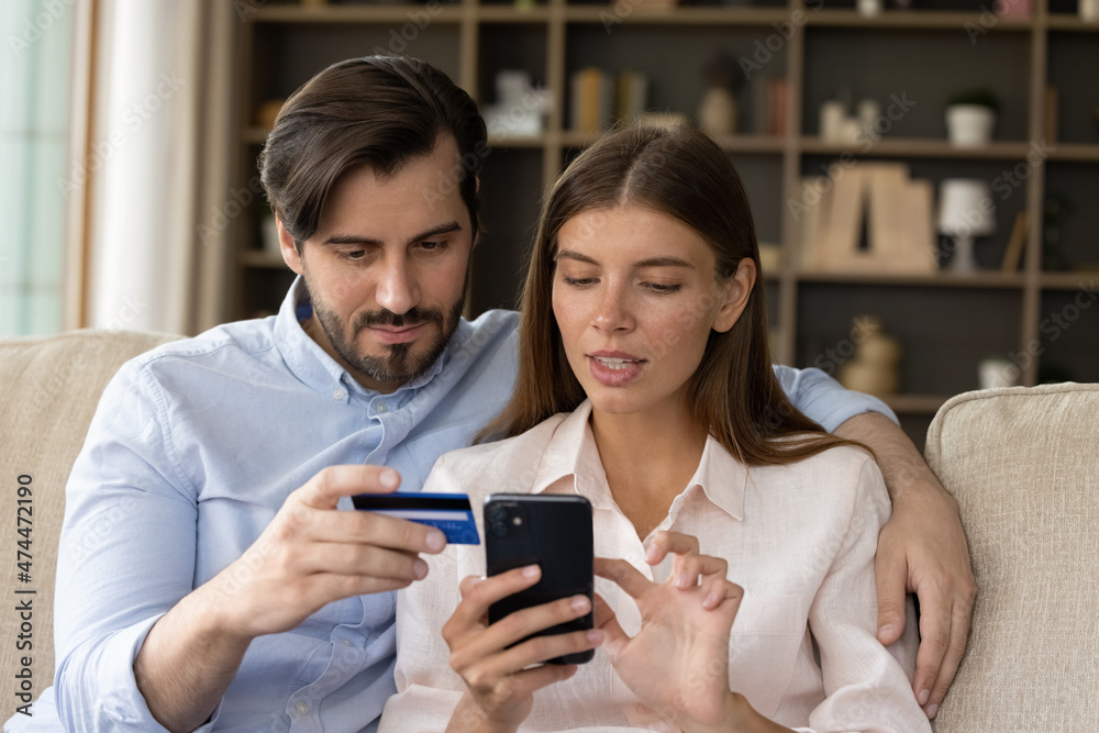 Focused millennial couple of consumers using banking app on mobile phone, credit card for online payment, transaction, transfer, money order, shopping on internet, paying bills, entering data Focused millennial couple of consumers using banking app on mobile phone, credit card for online payment, transaction, transfer, money order, shopping on internet, paying bills, entering data
