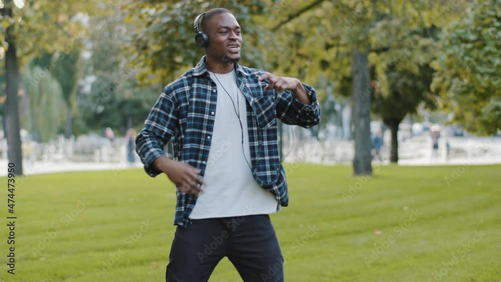 Young african american guy handsome man in headphones with mobile phone listening to music online enjoying audio song service sound app dancing in park outdoors on green grass moving rhythm movement Young african american guy handsome man in headphones with mobile phone listening to music online enjoying audio song service sound app dancing in park outdoors on green grass moving rhythm movement