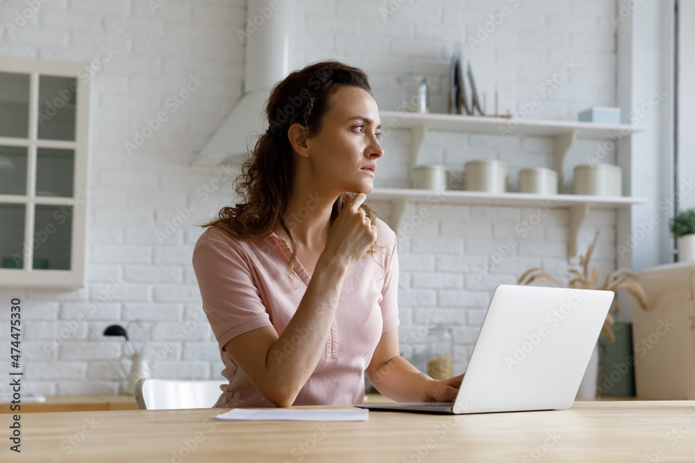 Serious thoughtful freelance employee woman working at laptop from home, using computer at kitchen table, thinking over online project future vision. Homeowner paying domestic bills on internet Serious thoughtful freelance employee woman working at laptop from home, using computer at kitchen table, thinking over online project future vision. Homeowner paying domestic bills on internet