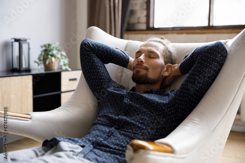 Calm happy millennial guy relaxing in pale comfortable armchair at home, resting with sleepy face, closed eyes, breathing fresh air deeply for stress reduce, calmness, serenity, meditation