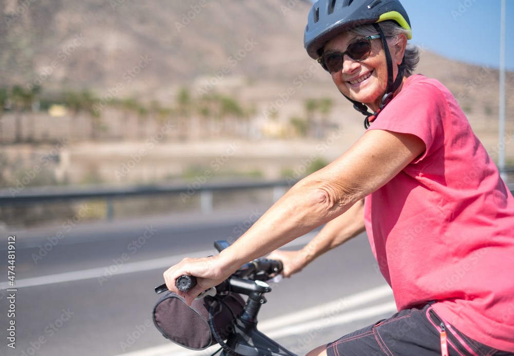 Portrait of happy mature woman, wearing protective helmet, on her bike in the sunny road. Healthy and active retirement concept Portrait of happy mature woman, wearing protective helmet, on her bike in the sunny road. Healthy and active retirement concept