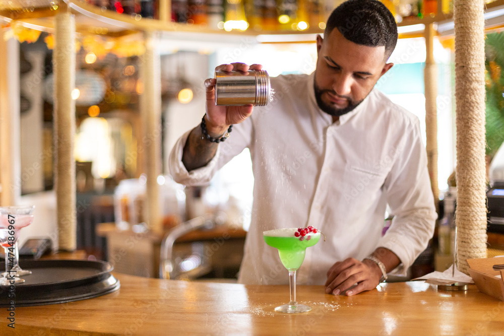 Young latin man carefully preparing an alcoholic drink in a modern cocktail bar. Space for text. Selective focus.