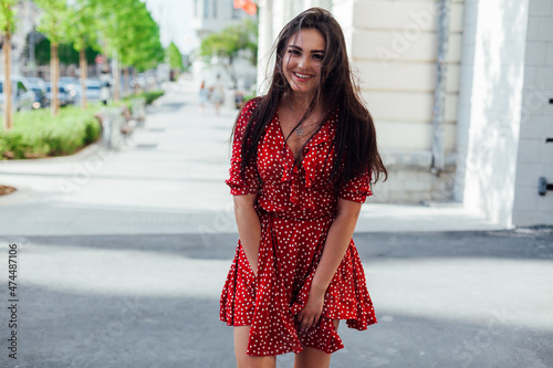 beautiful brunette woman in a red summer dress walks down the street © dmitriisimakov