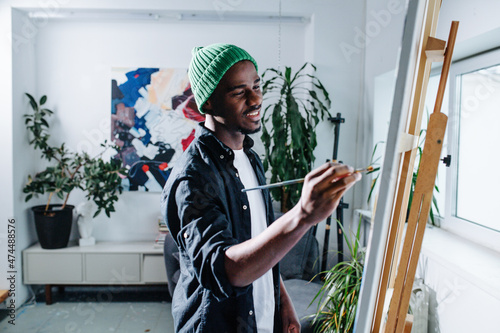 Fotografiet Inspired happy black man painting on an easel inside of his apartment