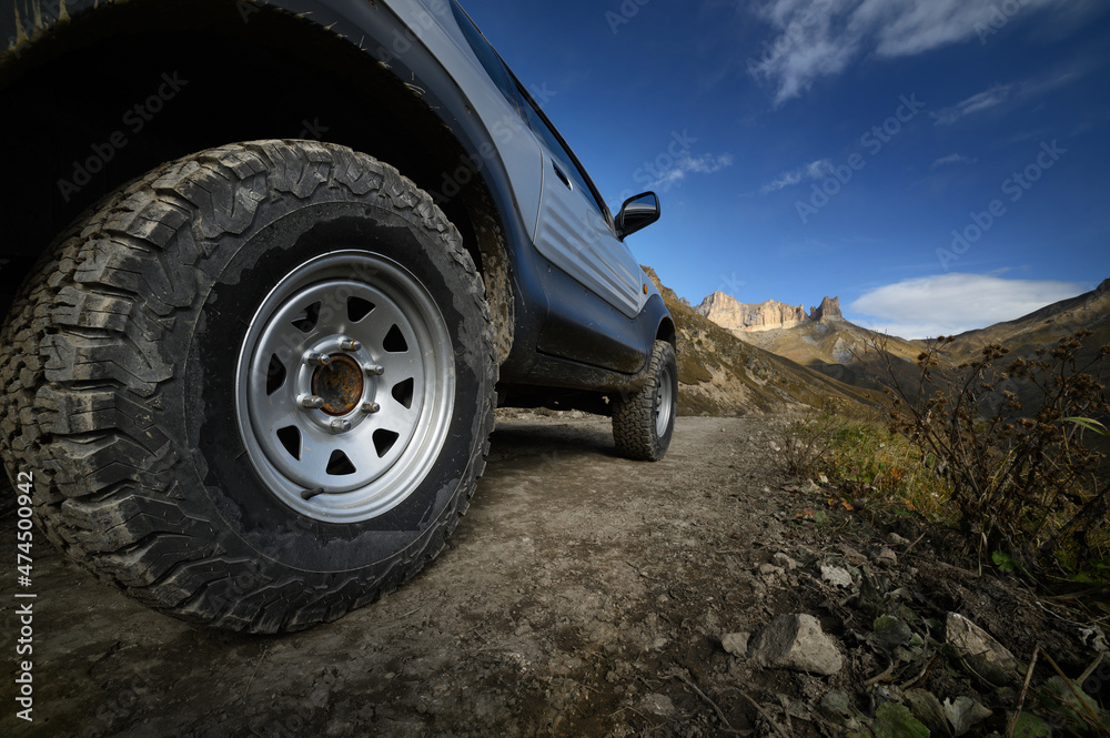 A large SUV car wheel with a large mud tread stands on a rocky road against the backdrop of mountains. Travel by off-road vehicles. Self-guided tourism by private car. A large SUV car wheel with a large mud tread stands on a rocky road against the backdrop of mountains. Travel by off-road vehicles. Self-guided tourism by private car.